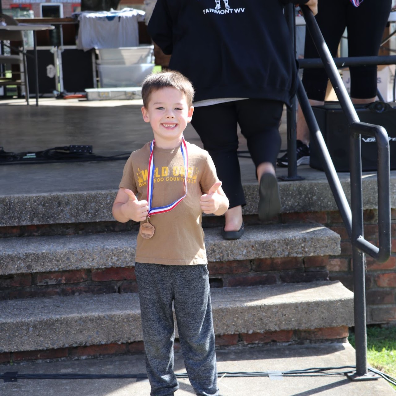 A young child stands proudly with a medal around his neck, giving a thumbs-up on a stage with steps and a blurred background.
