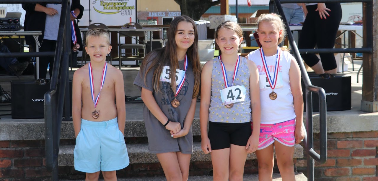 Four children proudly display their medals, standing on steps at an outdoor event, with a stage and spectators in the background.