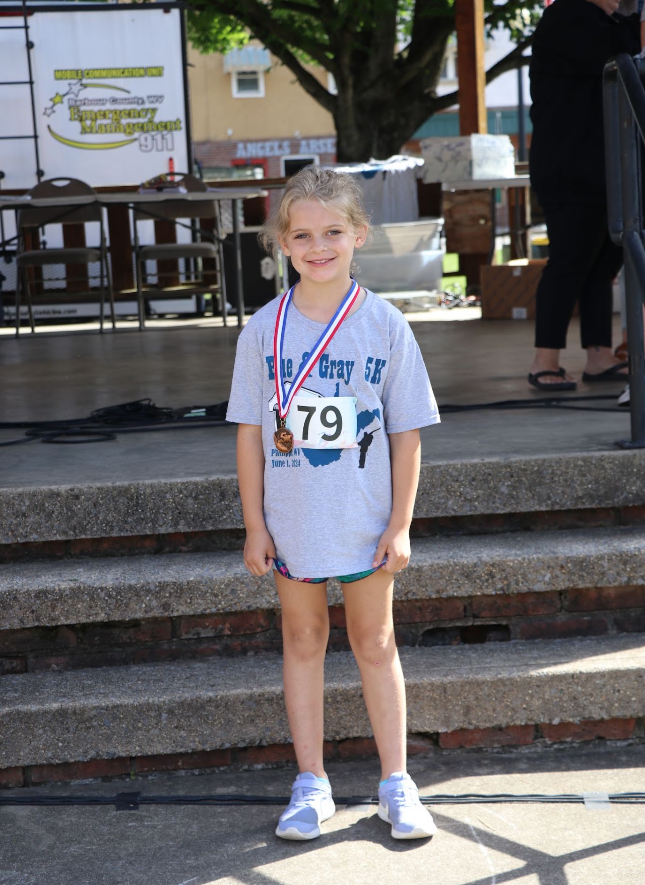 A child stands proudly on stage wearing a gray T-shirt, a medal, and race number 79, celebrating participation in a 5K event.