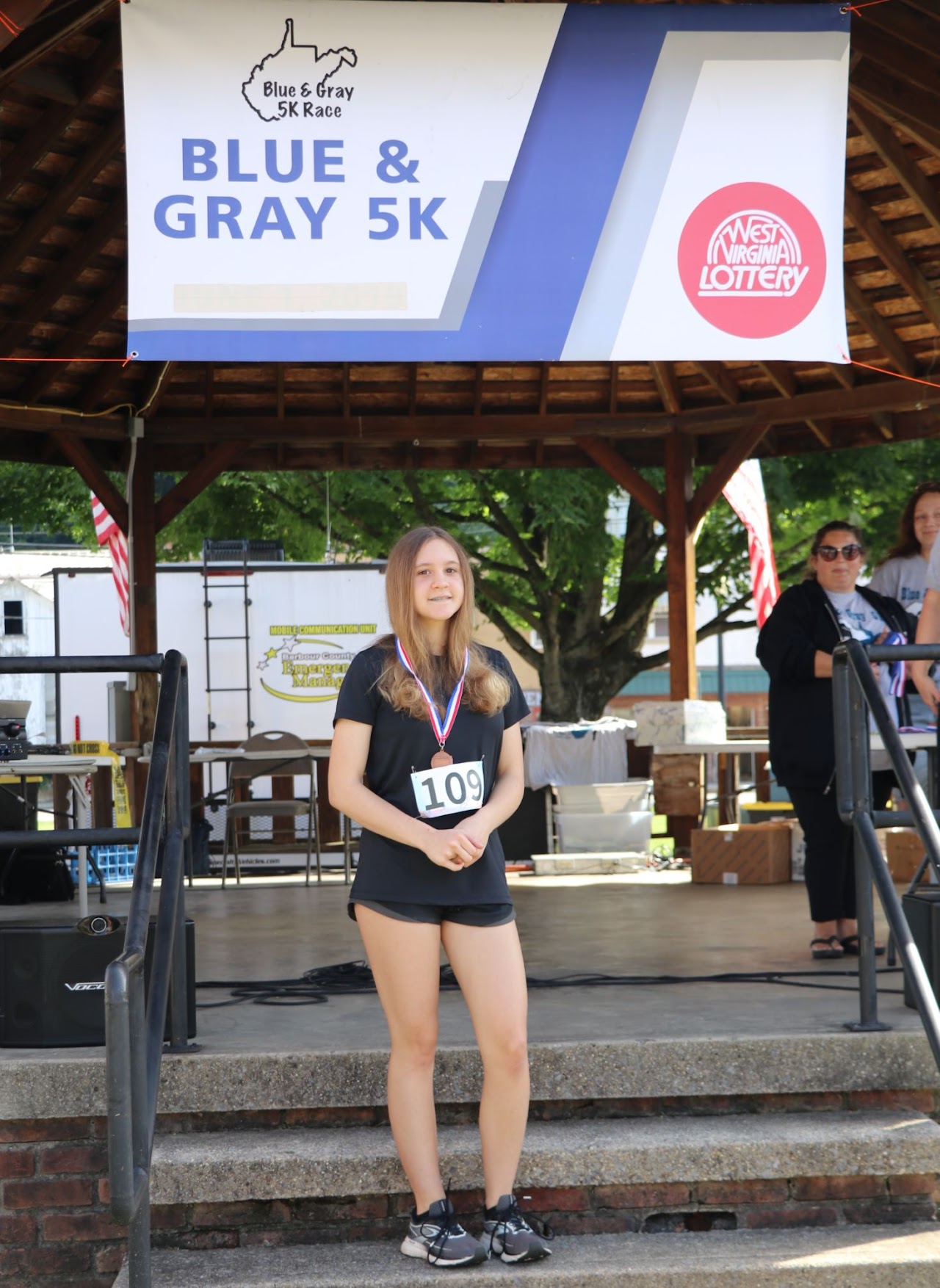 A participant at the Blue & Gray 5K race stands proudly on stage with a medal and race number displayed, under a colorful banner.