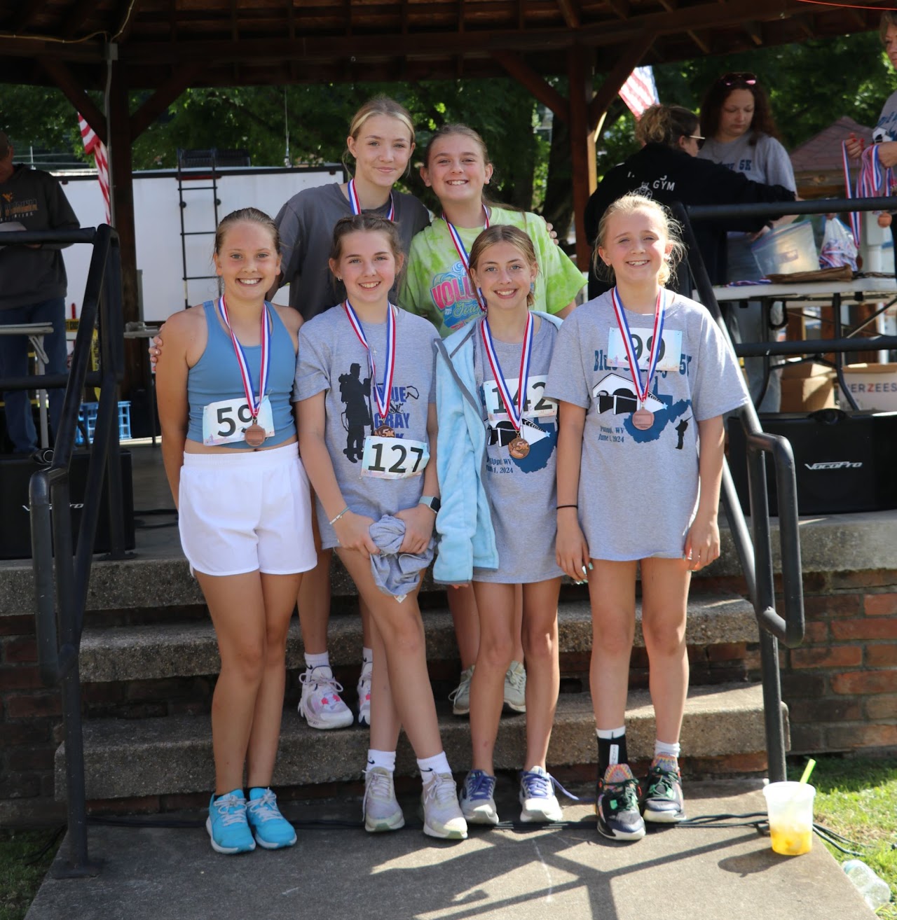 A group of five smiling young runners, wearing medals and race numbers, standing together at a competition event.