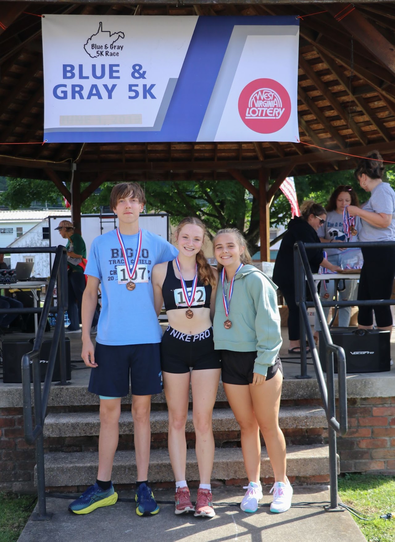 Three young athletes proudly display their medals after the Blue & Gray 5K race, standing under a festive banner.