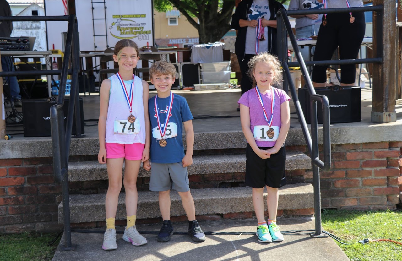 Three children stand proudly on a podium wearing medals after participating in a race, showcasing their numbers and achievements.