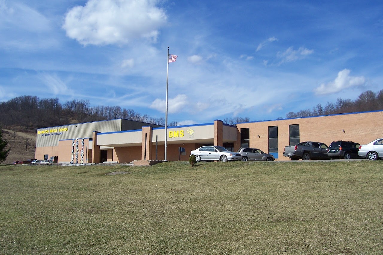 Exterior view of Belington Middle School, featuring a brick building with blue accents, flagpole, and parked cars on a grassy hillside.