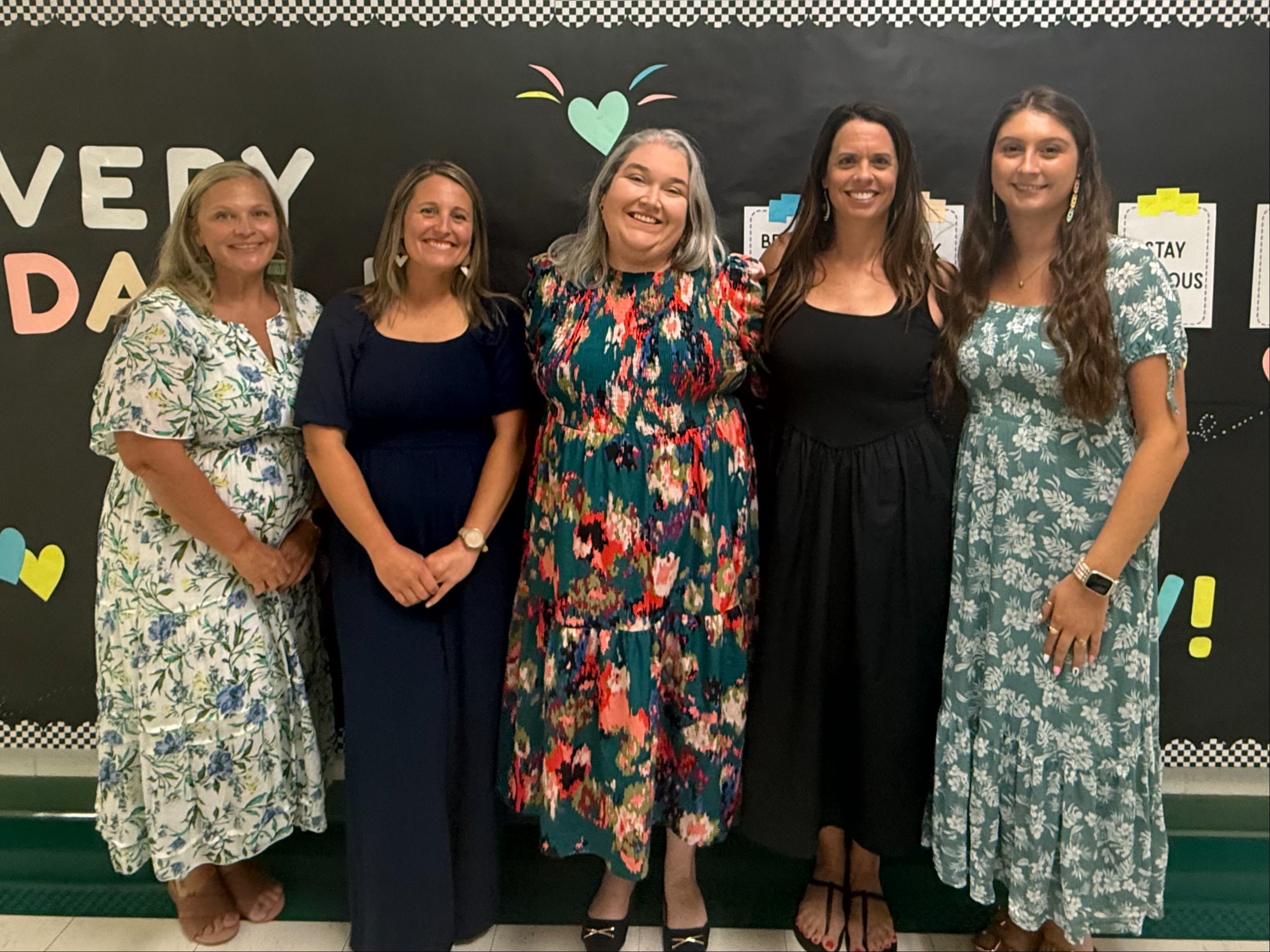 Five women standing side-by-side, smiling in front of a classroom bulletin board with colorful lettering and decorations.