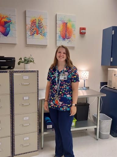 A smiling woman in colorful scrubs stands in a medical office, with colorful anatomical art on the walls and various office furniture around her.