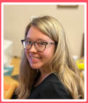 A woman with long, wavy blonde hair and glasses smiles while sitting at a desk with various items in the background.