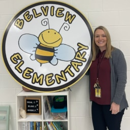 A woman stands next to a large circular sign that reads 'Belview Elementary' with a cartoon bee illustration.