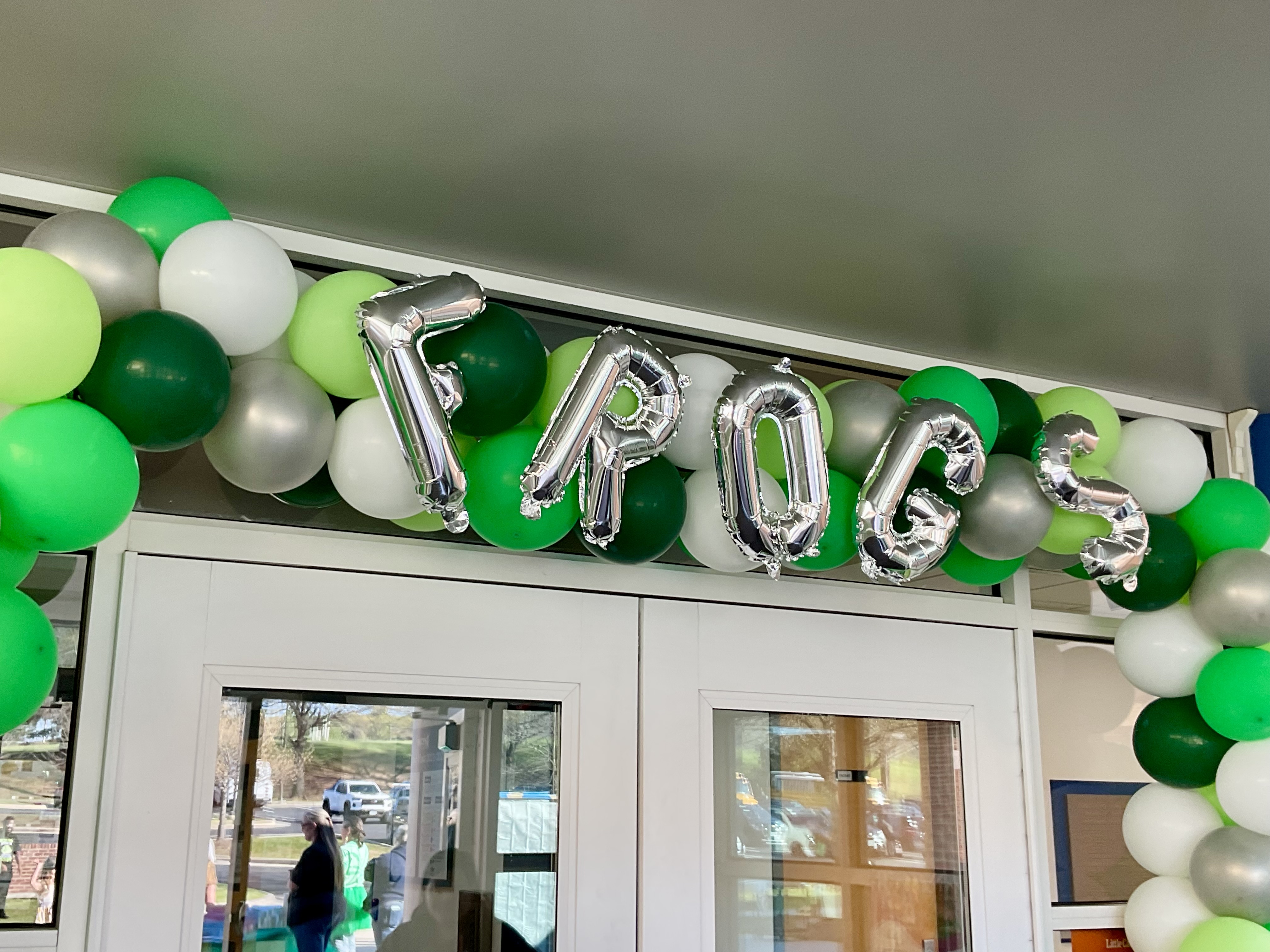 A festive balloon arch of green, white, and silver balloons features silver foil letter balloons that spell out the word "FROGS" above a doorway.