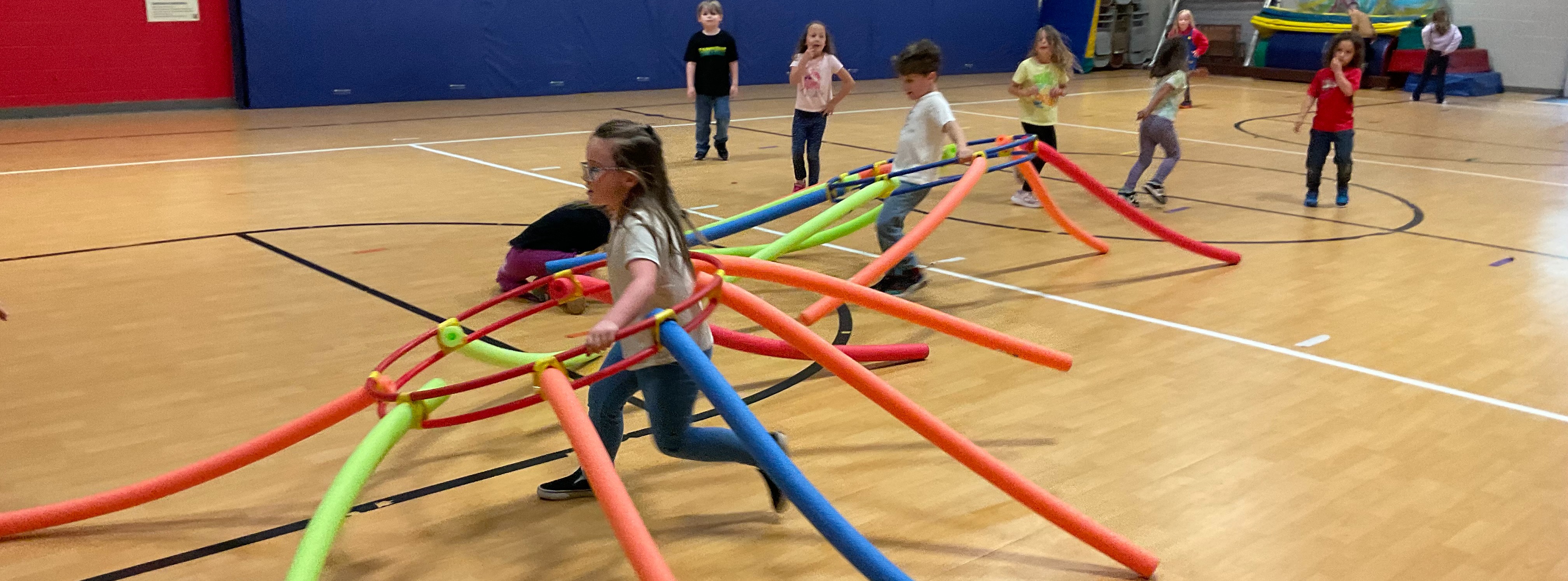 A class of children playing in gym. One child is carrying pool noodles shaped like an octopus and the other students are running and pretending to be crabs.