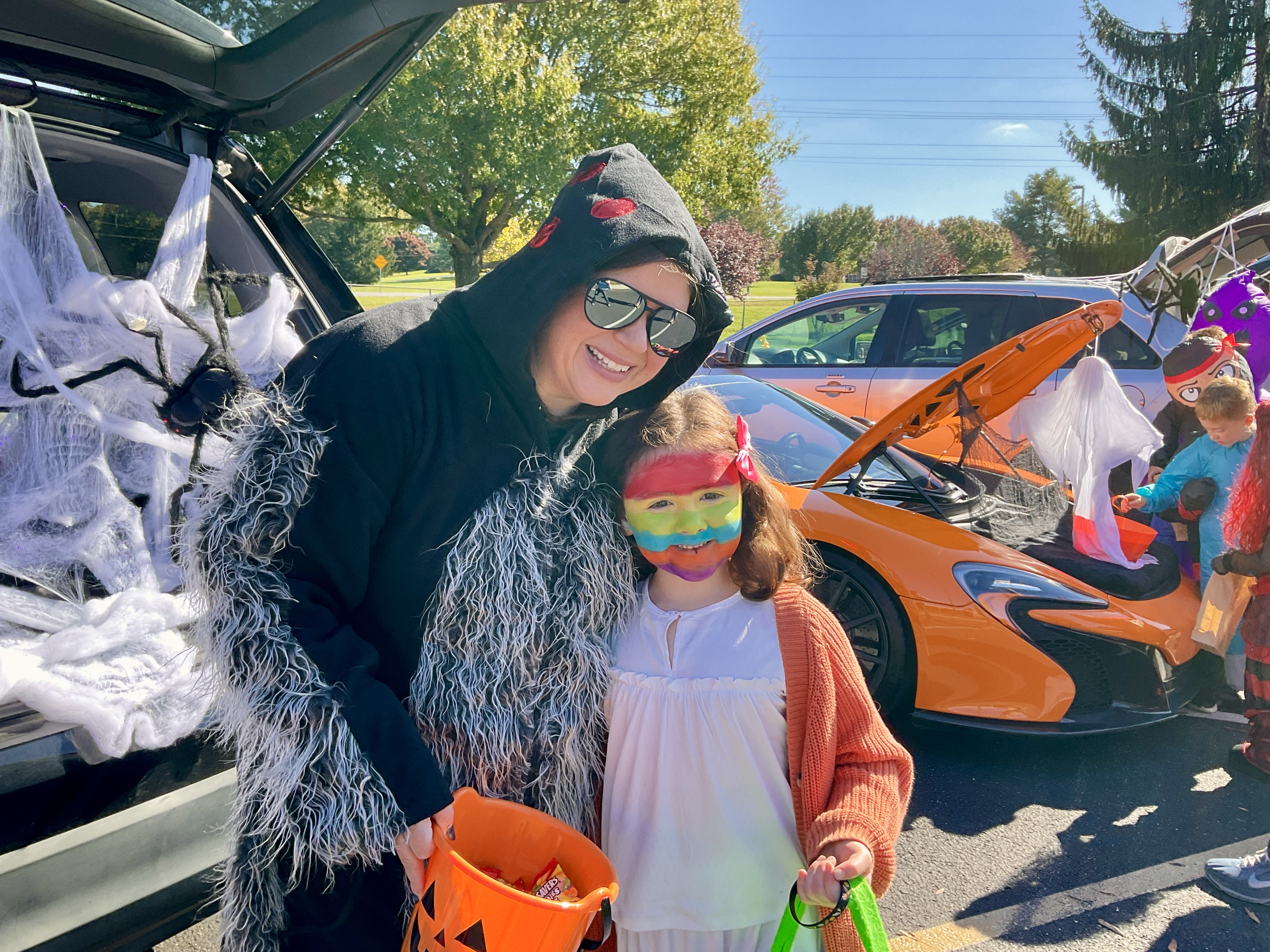 An adult in a furry black costume holding a treat bucket poses next to a young child with rainbow face paint in front of spiderweb-decorated car trunks.