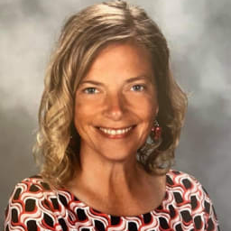A smiling woman with wavy blonde hair, wearing a patterned dress with red, black, and white colors, poses against a soft gray background.