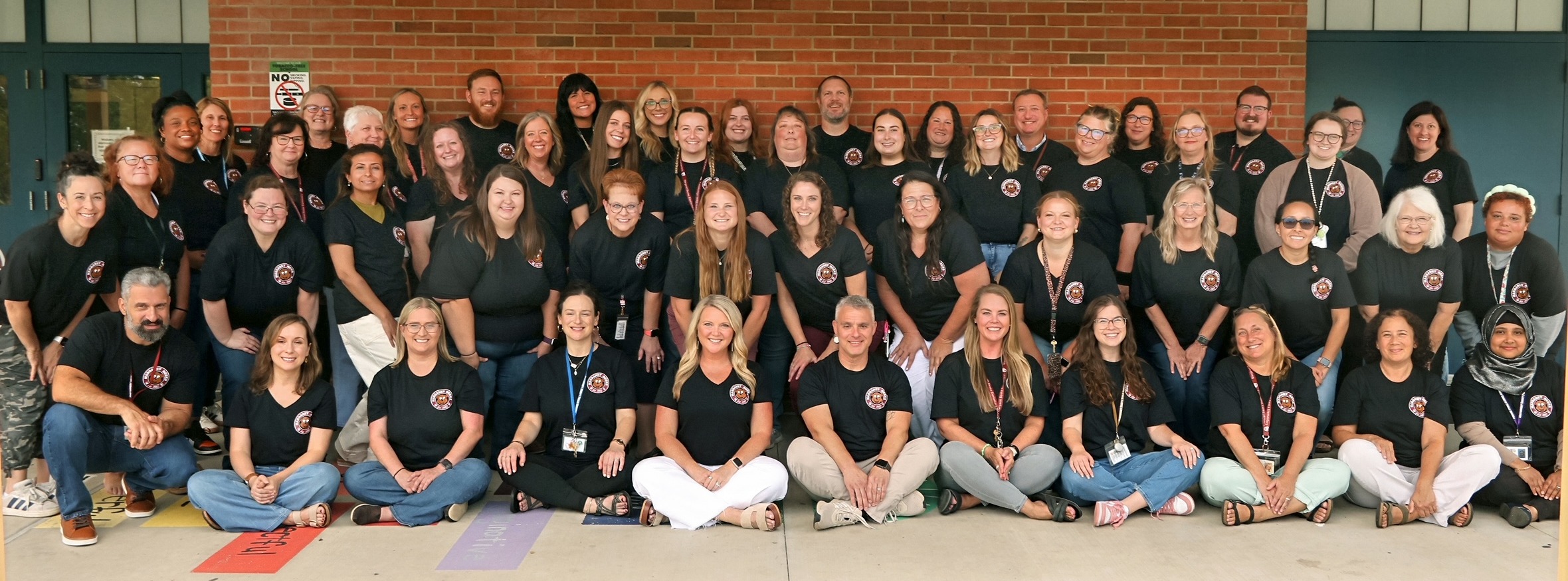 A large group portrait of school staff and teachers standing and sitting together outside in front of a red brick wall. Most are wearing matching black t-shirts with a circular school logo.