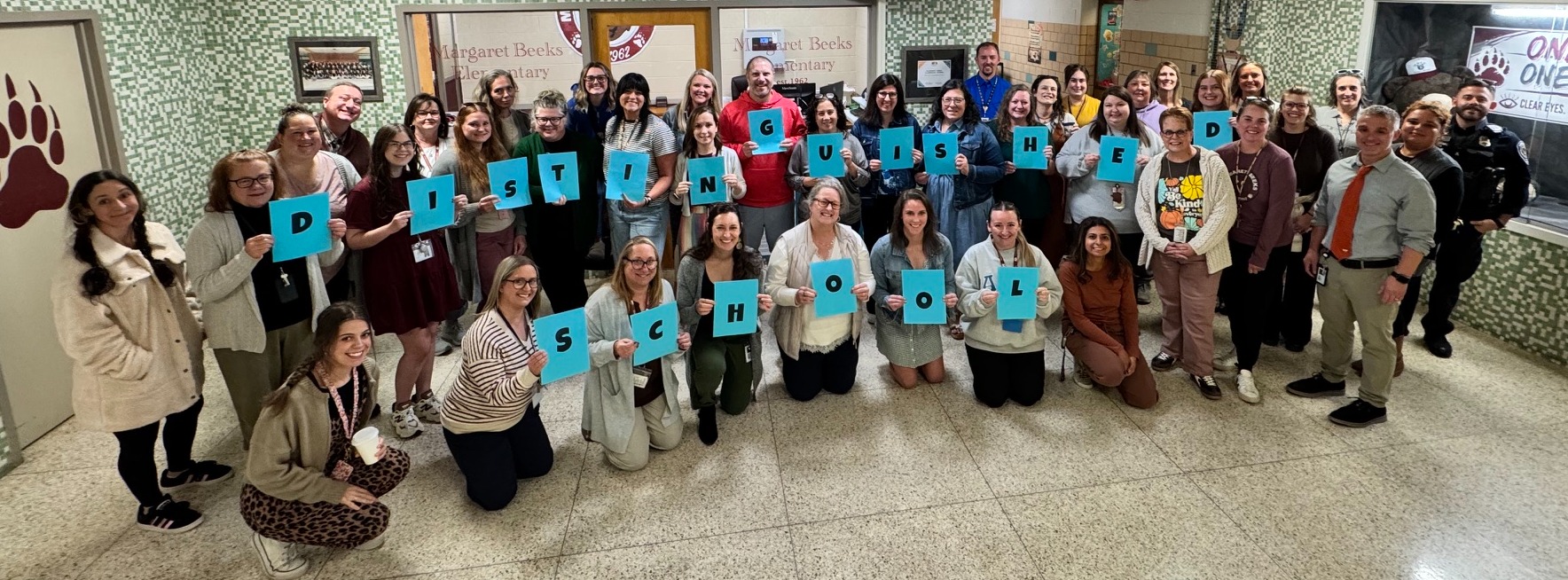 A group of staff members inside a school hallway posing for a picture, with several individuals holding up individual blue signs that spell out "DISTINGUISHED SCHOOL." On the left wall, a large paw print icon is visible, and in the background, text reads "Margaret Beeks Elementary" and "ONE TEAM ONE PURPOSE".