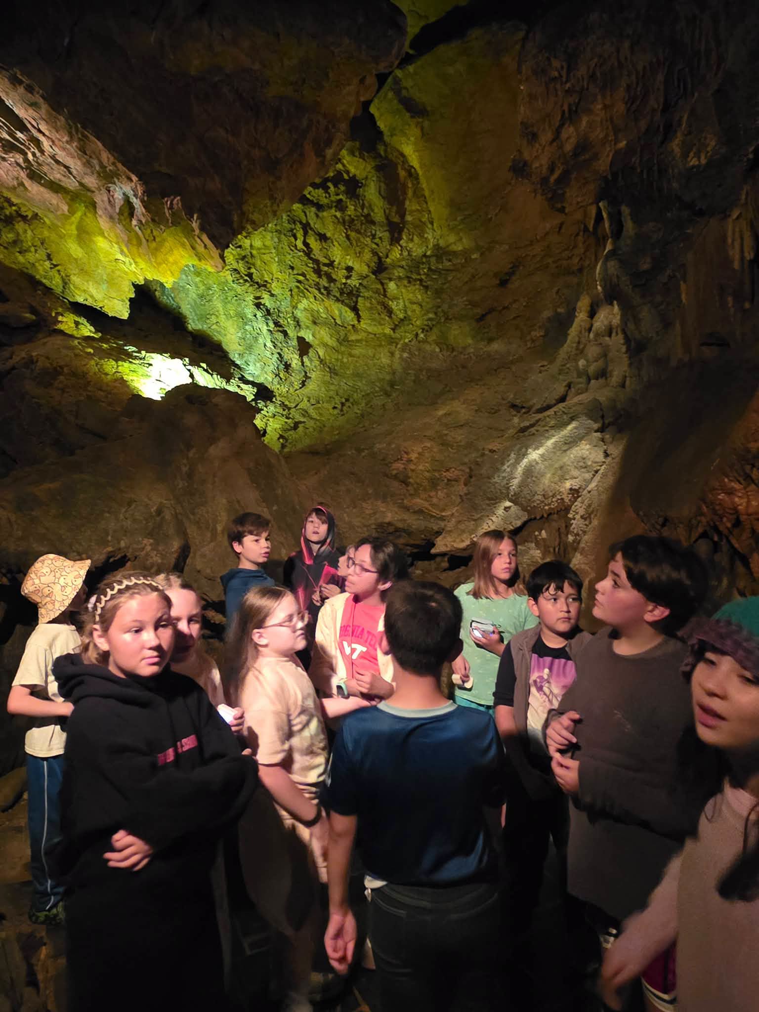A group of middle school students gathered inside a dimly lit cave, looking around at the surrounding rock formations illuminated by soft yellow lighting overhead.