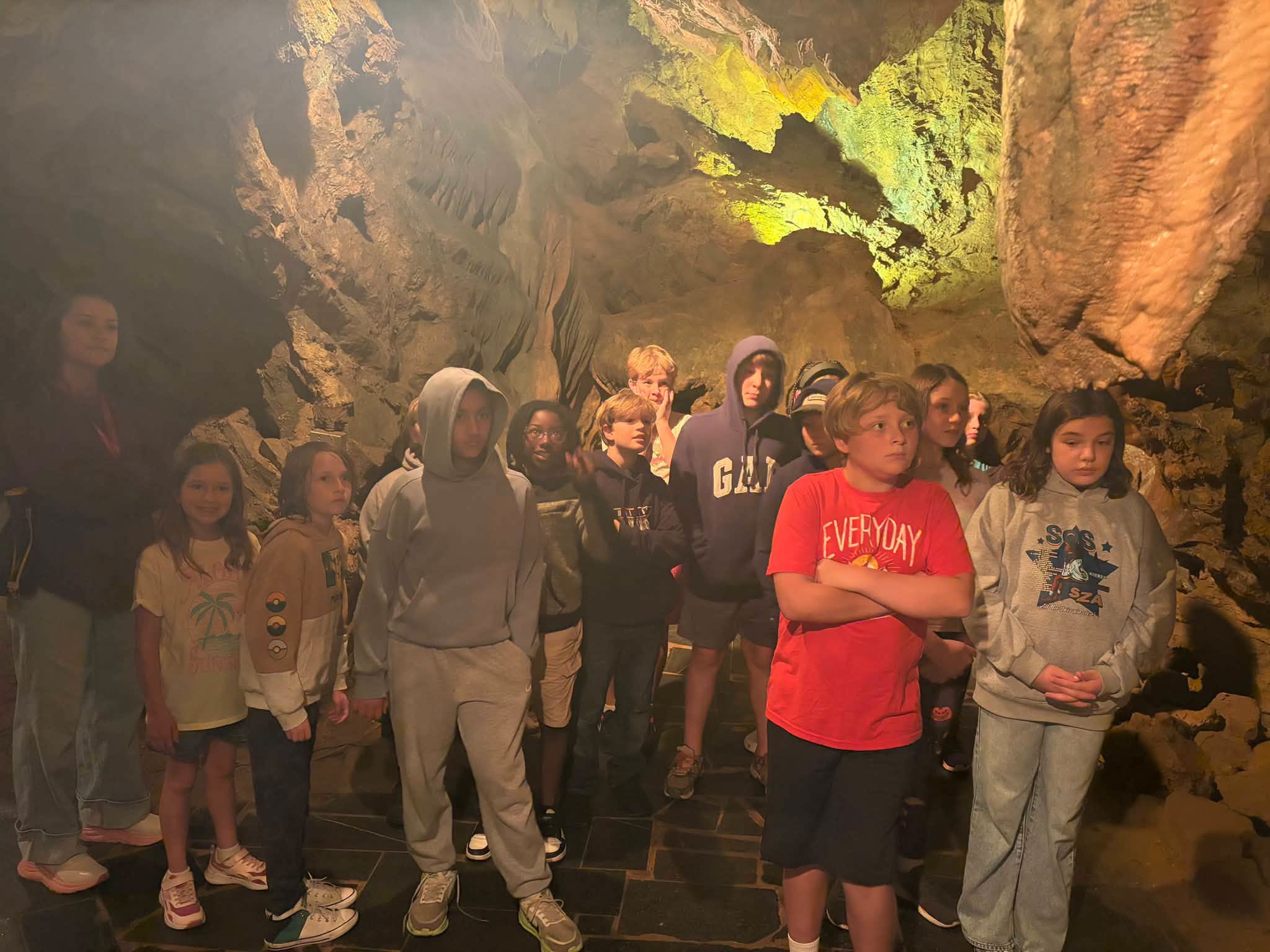 Students and a chaperone standing together on a stone pathway inside a large cavern, with textured rock walls and mineral deposits lit up behind them.
