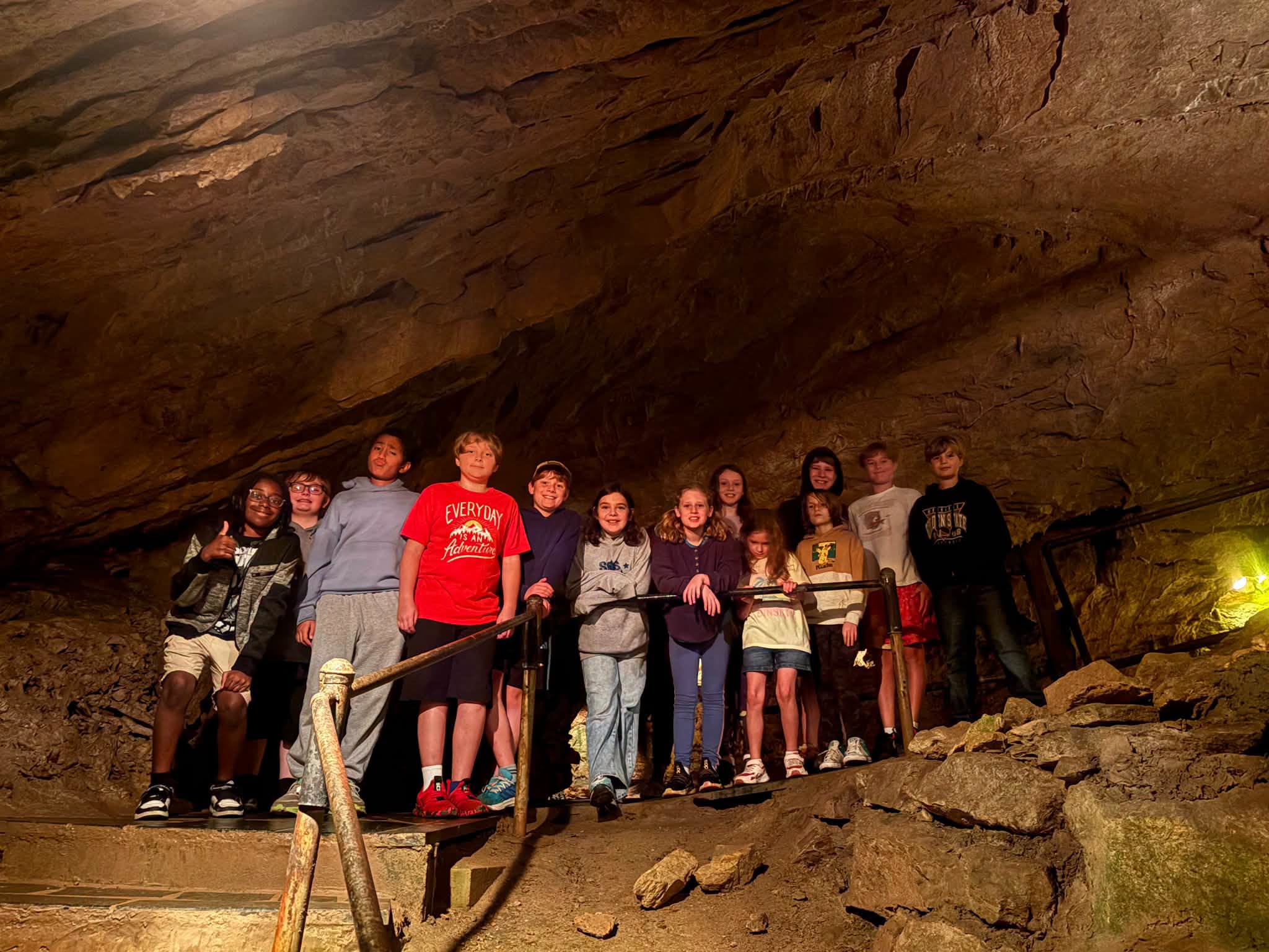  A group of students lining up along a metal handrail on a stone staircase inside a dimly lit cavern, smiling for a photo during a cave field trip.   