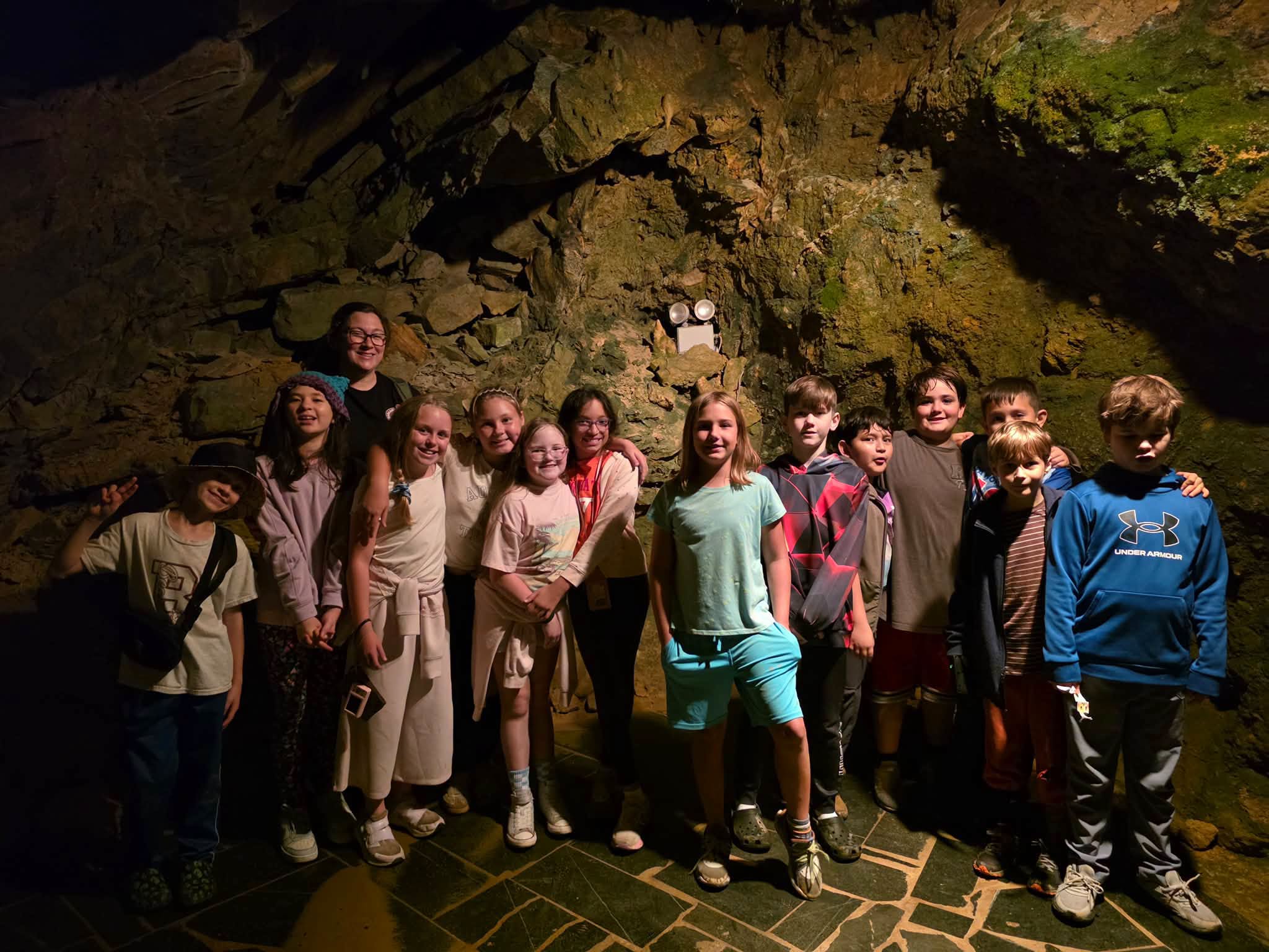 A group of students and their teacher posing together for a group photo inside an illuminated cavern in front of a natural stone wall.