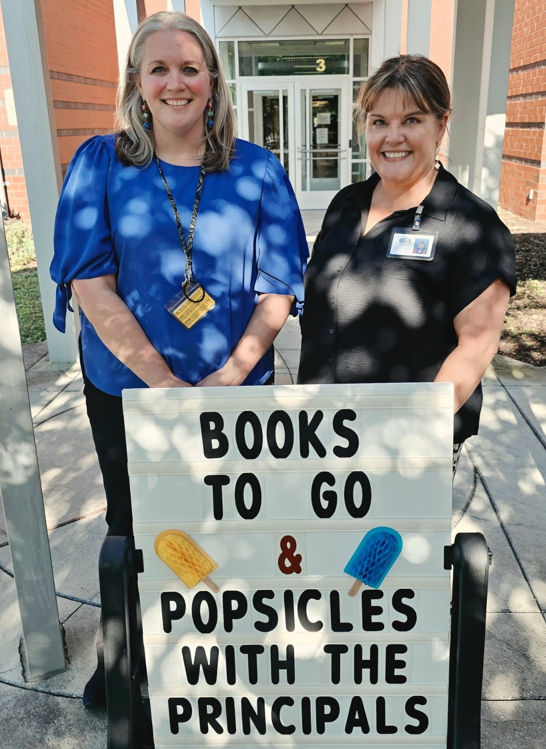 Two women standing behind a sign that says "Books to Go and Popsicles with the Principals"