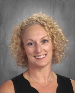 A woman with curly blonde hair smiles at the camera, wearing a black top, against a gray background.