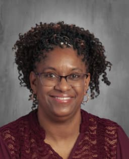 A smiling woman with curly hair, wearing glasses and a maroon top, poses for a portrait against a neutral background.