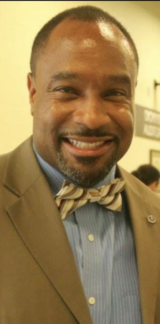 A smiling man wearing a beige suit jacket and a bow tie, standing in a well-lit indoor setting.