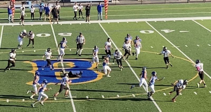 A high school football game in progress, with players in black and white uniforms on a green field marked with a large blue letter 'B'.