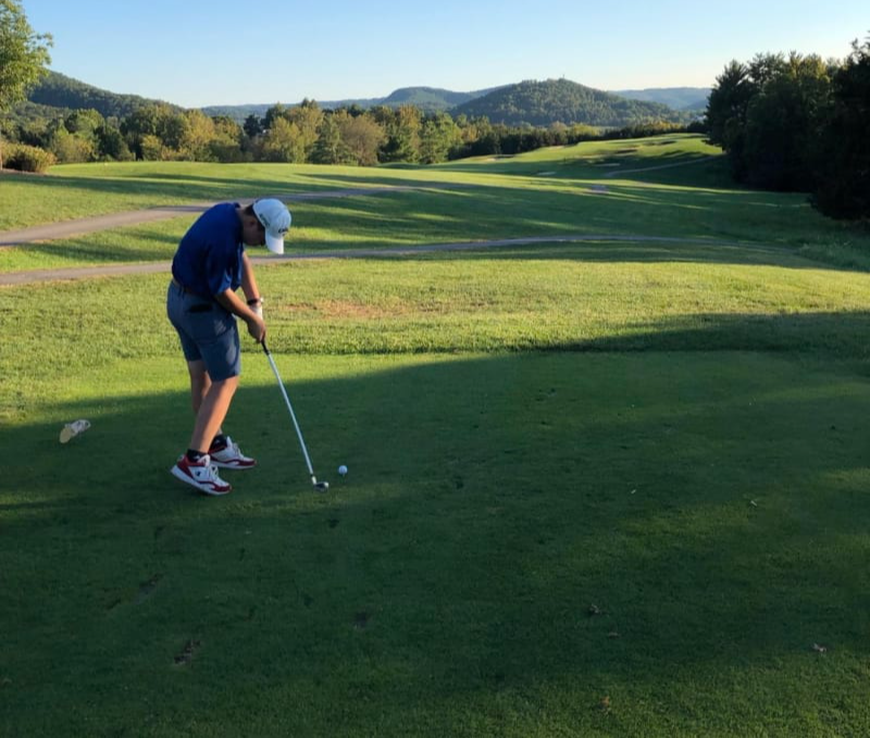 A young golfer prepares to take a shot on a lush green golf course with rolling hills and a clear blue sky in the background.