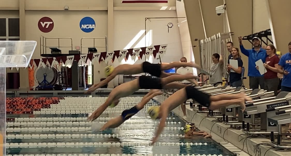 A sequence of swimmers diving into a pool from starting blocks during a competition, with spectators and officials in the background.