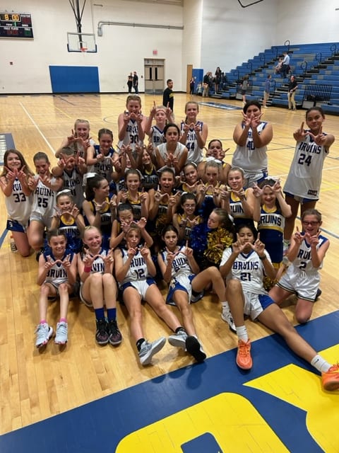 A large group of young female basketball players poses together on a gymnasium floor, celebrating with their hands raised in a victory gesture, surrounded by basketballs and wearing team jerseys.