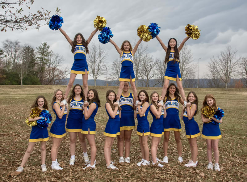 A cheerleading squad poses outdoors, with some members performing stunts and holding pom-poms, while others stand in a pyramid formation on the ground.
