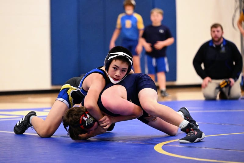 Two young wrestlers are engaged in a match on a blue wrestling mat, with spectators and coaches visible in the background.