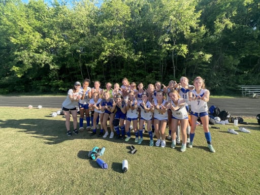 A group of young female soccer players poses together on a grassy field, smiling and celebrating with their arms raised, surrounded by trees in the background.