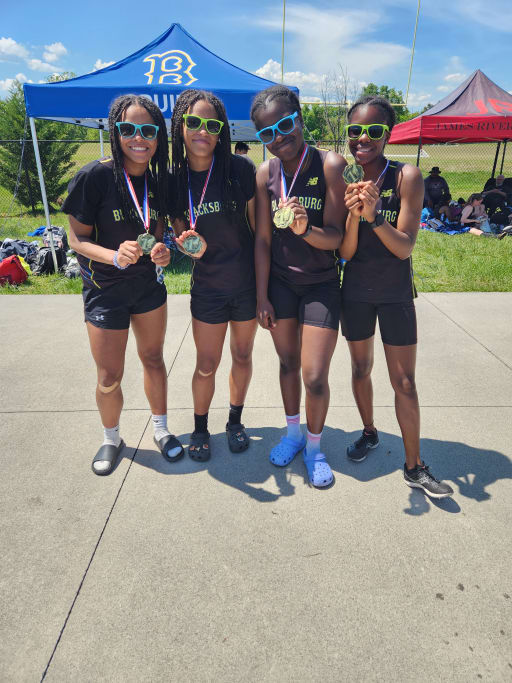 Four athletes wearing black uniforms and sunglasses proudly display their medals while standing outdoors under a blue sky.
