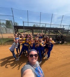 A group photo of a softball team, consisting of players and coaches, all wearing matching black shirts with blue accents, standing on a softball field with a cloudy sky in the background.