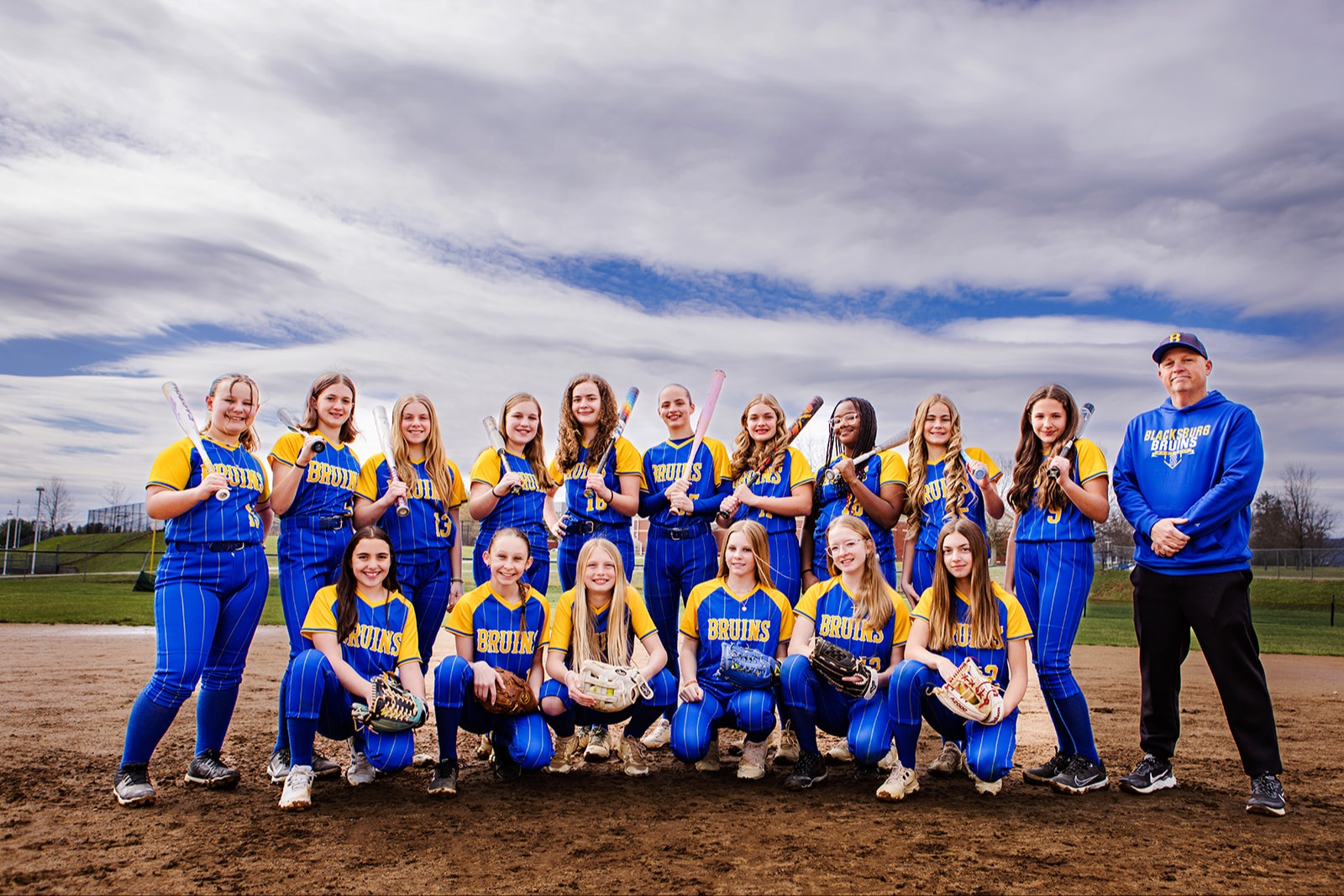 A group photo of a softball team, consisting of players and coaches, all wearing matching black shirts with blue accents, standing on a softball field with a cloudy sky in the background.