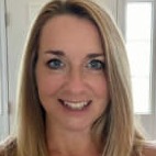A woman with long blonde hair smiles at the camera, standing in a well-lit room with white doors in the background.