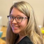 A woman with long, wavy blonde hair and glasses smiles while sitting at a desk with various items in the background.