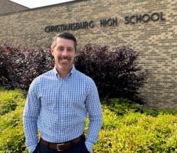 A man stands smiling in front of Christiansburg High School, with greenery and a brick wall in the background.