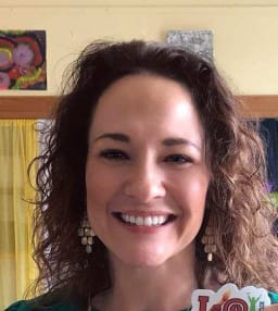 A smiling woman with curly hair, wearing earrings, stands in front of a colorful background with artwork.