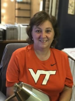A woman with short brown hair is smiling while wearing an orange Virginia Tech t-shirt, seated at a desk with a lamp and office supplies in the background.