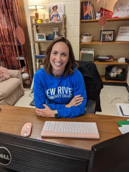 A smiling woman with shoulder-length brown hair sits at a desk, wearing a blue sweatshirt with 'New River Community College' printed on it, with a computer keyboard and mouse in front of her.