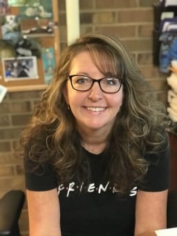 A smiling woman with curly hair and glasses is sitting at a desk, wearing a black t-shirt that says 'FRIENDS'.