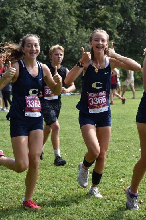Two female runners in athletic uniforms give thumbs up while running on a grassy field, with other runners visible in the background.