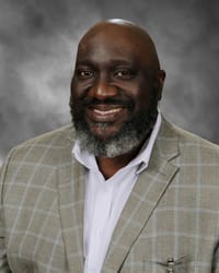 A smiling man with a beard, wearing a light-colored plaid blazer over a white shirt, poses against a blurred gray background.