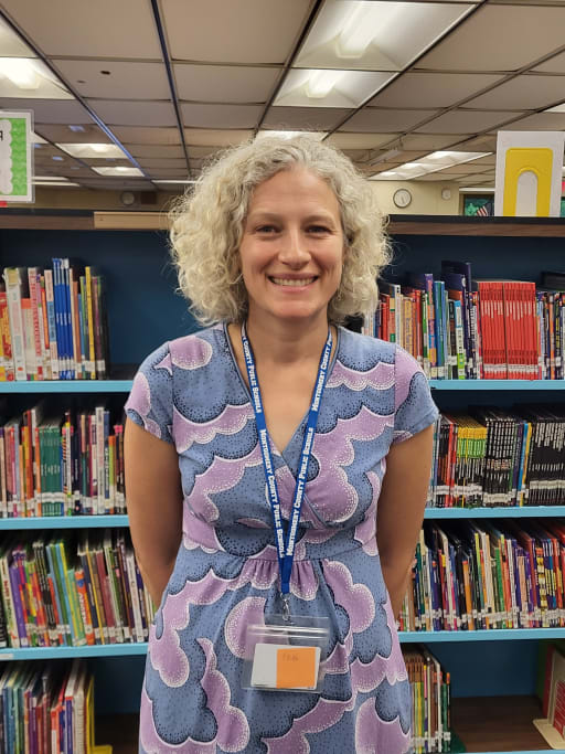 A smiling woman with curly hair stands in front of a bookshelf filled with colorful children's books, wearing a patterned dress and a name tag.