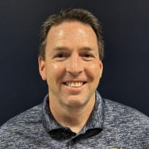 A smiling man wearing a dark blue polo shirt with a logo on the left side, standing in front of a dark background.