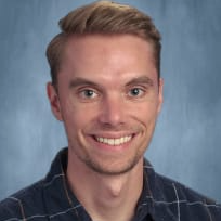 A smiling man with short blonde hair, wearing a dark plaid shirt, poses against a blue background.