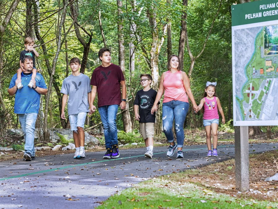 1. A family strolls along a scenic trail, passing a sign that marks their path through nature.   2. A family enjoys a walk on a trail, with a directional sign visible alongside them in the natural surroundings.  3. A family walks together on a trail, accompanied by a sign that provides information about the route ahead.