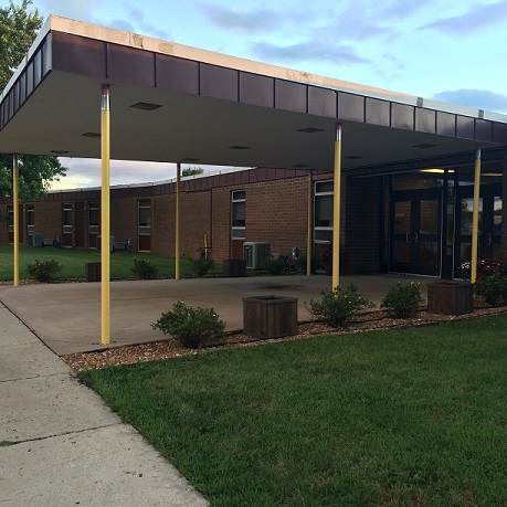 A view of a school entrance featuring a covered walkway supported by yellow pillars, with landscaped areas and a grassy lawn in the foreground.